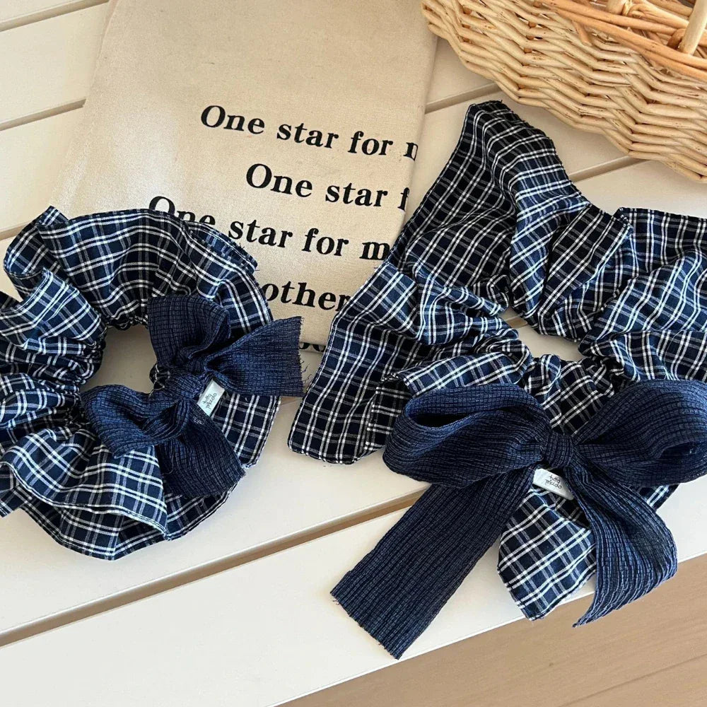 Two scrunchies with navy and white plaid patterns and dark blue textured bows are on a white table next to a canvas bag with text.