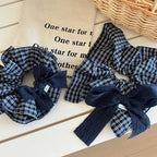 Two scrunchies with navy and white plaid patterns and dark blue textured bows are on a white table next to a canvas bag with text.