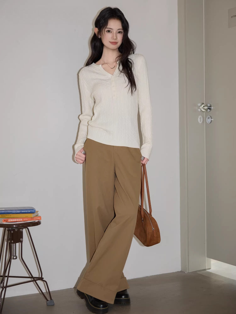 Woman standing next to a stack of books and a stool, wearing a white cable knit henley sweater and brown wide-leg pants, holding a brown handbag.