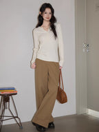 Woman standing next to a stack of books and a stool, wearing a white cable knit henley sweater and brown wide-leg pants, holding a brown handbag.