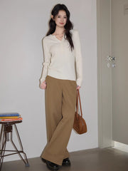 Woman standing next to a stack of books and a stool, wearing a white cable knit henley sweater and brown wide-leg pants, holding a brown handbag.
