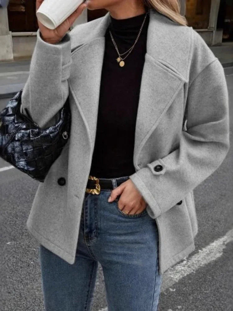 Close-up of a woman's torso wearing a grey peacoat, black turtleneck, and gold necklaces.