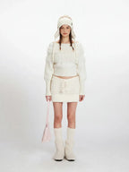 Model wearing a white off-the-shoulder sweater and matching mini skirt with a white knit hat, white knee-high socks, and white fuzzy boots. She is holding a pink handbag.