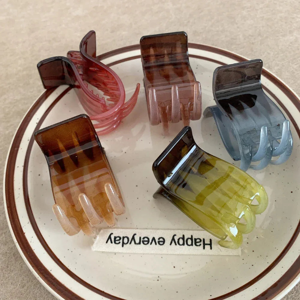 Five hand-shaped claw clips are displayed on a plate with a striped rim, featuring colors like brown, pink, green, and blue-grey.