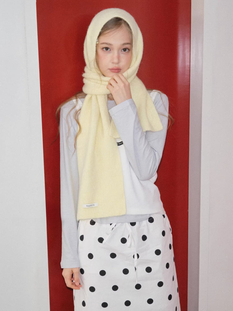 Model wearing a light yellow ribbed knit scarf wrapped around their head and neck, with hands touching the scarf near their chin.