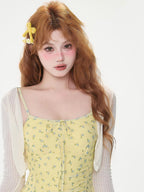 Close-up of a woman's face and upper body, wearing a yellow floral print mini dress with a white sheer cardigan