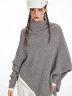 Close-up of a woman wearing a grey asymmetrical knit poncho with a turtleneck, looking at the camera