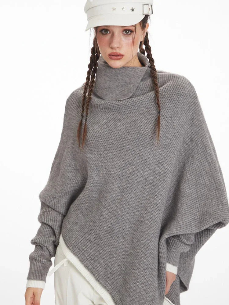 Close-up of a woman wearing a grey asymmetrical knit poncho with a turtleneck, looking at the camera