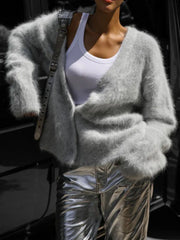 Model wearing a light grey fuzzy V-neck button-up cardigan over a white tank top and silver metallic pants, leaning against a vehicle.