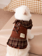 The back of a white cat wearing a brown plaid dress with bows and a pleated skirt.