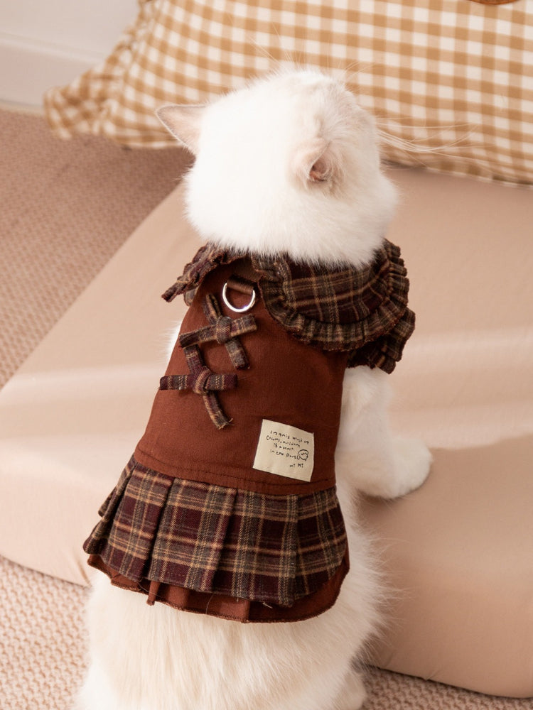 The back of a white cat wearing a brown plaid dress with bows and a pleated skirt.