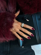 Close-up of a woman's hand with burgundy nails and rings, holding a black clutch bag next to a burgundy faux fur jacket