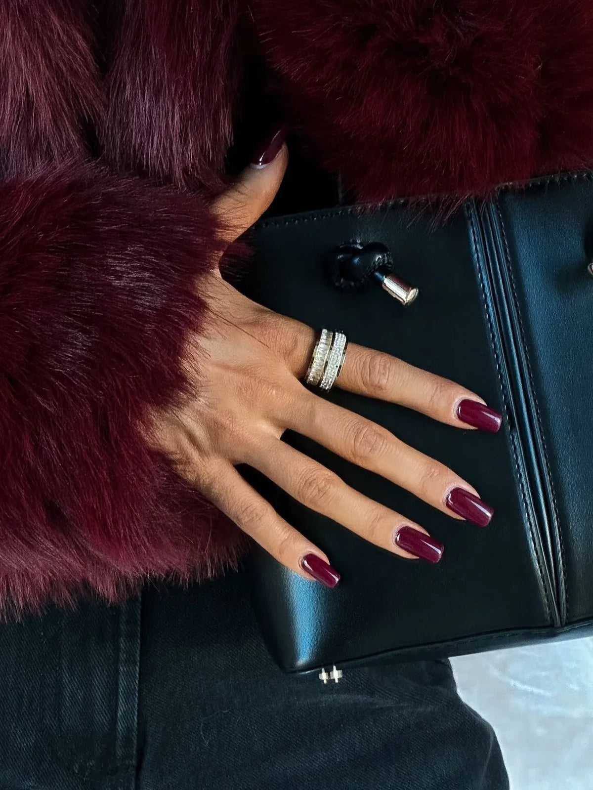 Close-up of a woman's hand with burgundy nails and rings, holding a black clutch bag next to a burgundy faux fur jacket