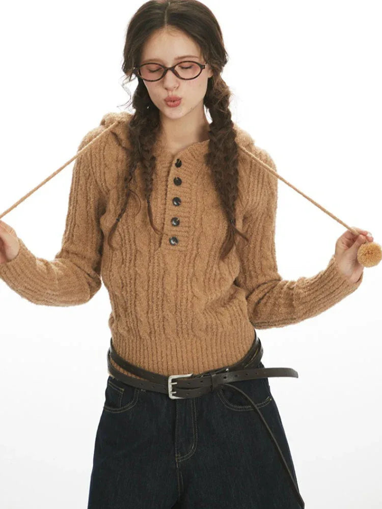 Close-up of a woman wearing a brown cable knit hooded sweater with buttons and pom-poms, holding the drawstring.