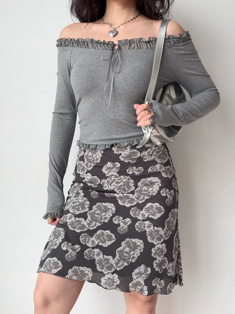 Woman wearing a grey off-the-shoulder ribbed knit top with ruffled trim and tie front, paired with a floral print skirt and a silver handbag.