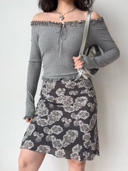 Woman wearing a grey off-the-shoulder ribbed knit top with ruffled trim and tie front, paired with a floral print skirt and a silver handbag.