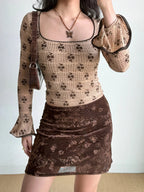 Full body shot of a person wearing a fitted, long-sleeved square neck top with an allover brown floral print on a beige textured knit fabric. The top has flared sleeves with dark scalloped trim. A dark brown velvet skirt with a floral pattern and a dark brown handbag are also visible.