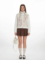 Woman wearing a floral print fleece bomber jacket with cable knit sleeves, brown pleated skirt, white knee-high socks, and black Mary Jane shoes, holding a white bucket bag.