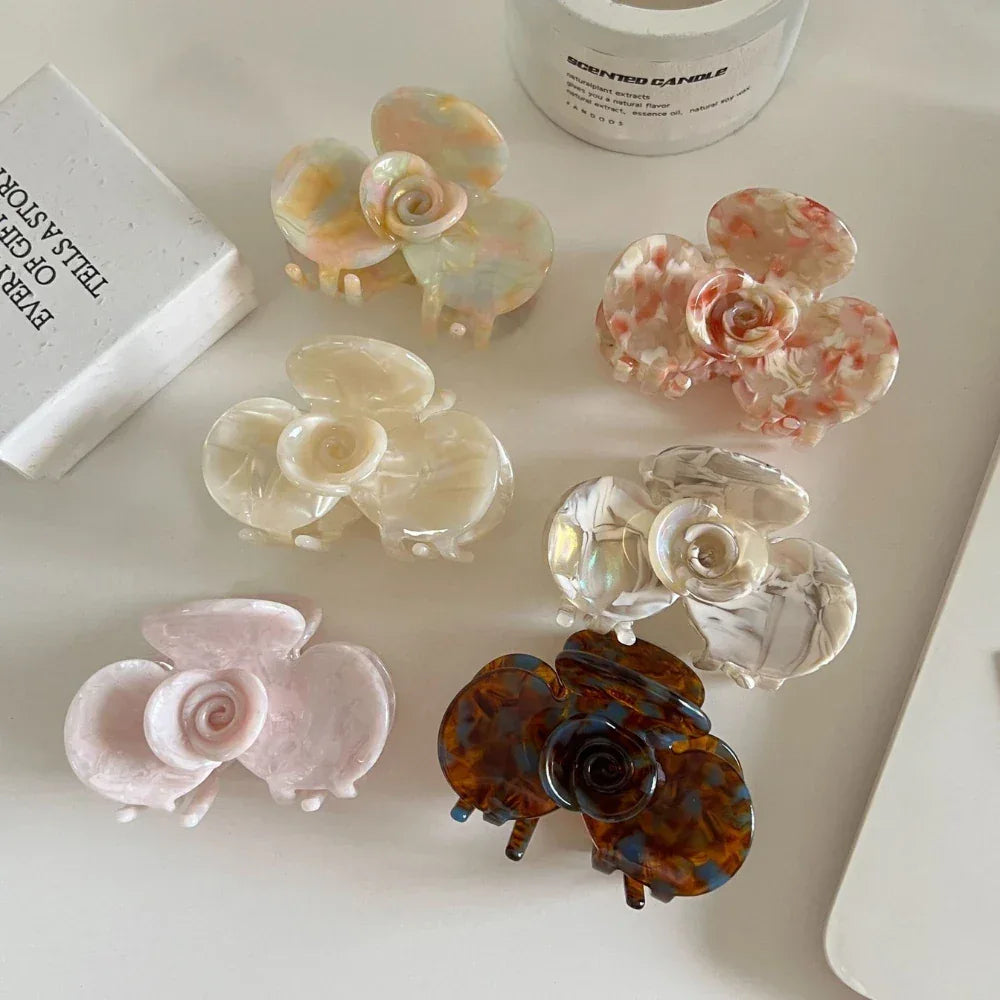 A top-down view of six floral-shaped acetate hair claw clips, showcasing a variety of colors and marbled patterns, placed on a white surface.