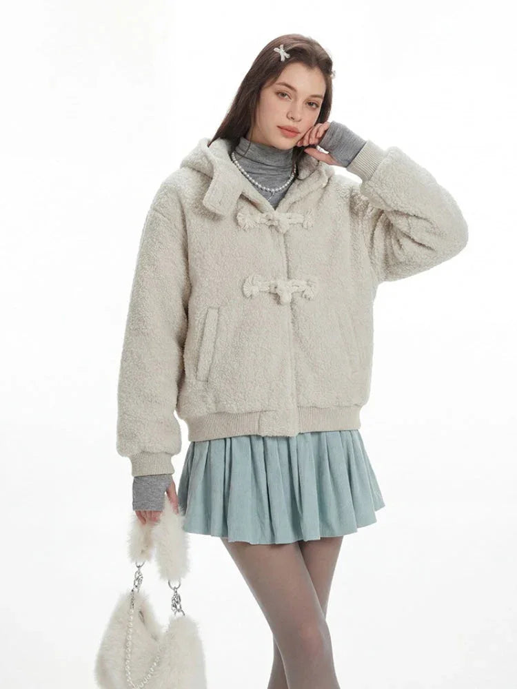Model posing with hand on chin, wearing a cream fuzzy hooded bomber jacket with toggle closure, a light blue pleated skirt, grey turtleneck, sheer tights, white leg warmers, and a white fuzzy handbag.