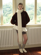 Woman wearing a brown velour sweatshirt with a white scarf, grey shorts, grey socks, and white sneakers, standing in front of a window.
