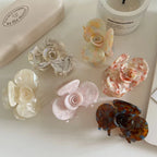 A flat lay of six floral-shaped acetate hair claw clips in different pastel and marbled designs, arranged on a white surface with a candle and box.