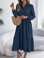 Woman standing holding a straw bag and wearing a long-sleeved blue midi dress with a collared neckline and tiered hem