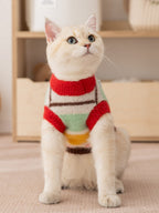 A cream-colored cat wearing a colorful striped sweater with red trim sits facing forward.
