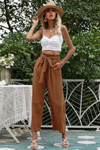 Woman wearing a white crop top and brown wide-leg pants with a tie belt, wearing a straw hat.
