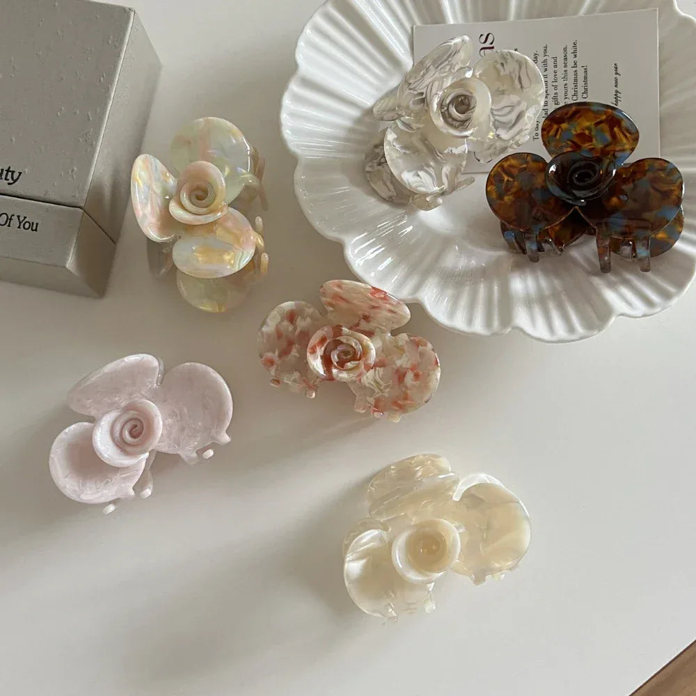 A top-down view of several floral-shaped acetate hair claw clips, including a brown and blue one, on a white surface with decorative elements.
