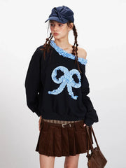 Close-up of the front of the black sweatshirt, highlighting the off-the-shoulder design with ruffled trim and the textured appliqué bow.
