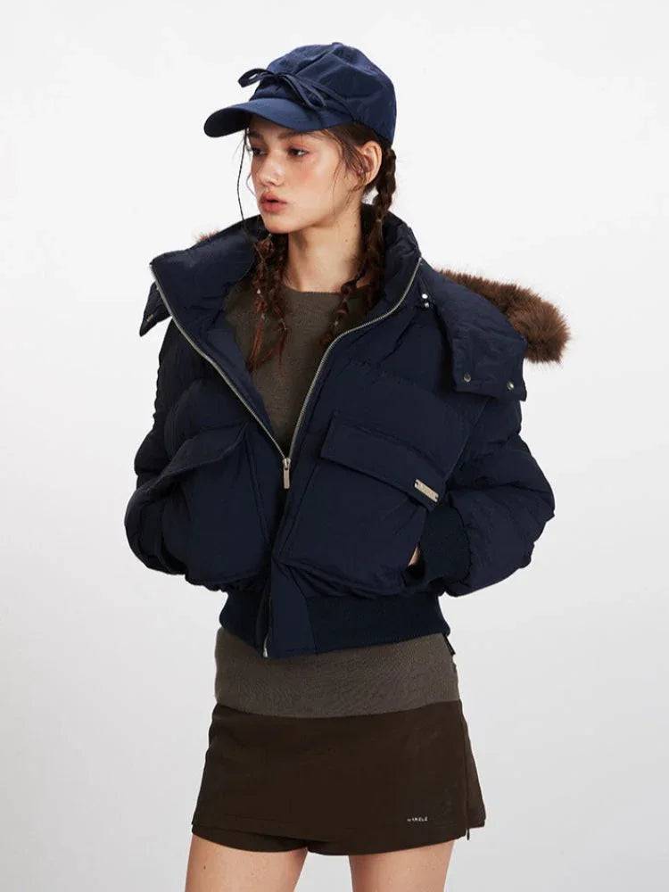 Close-up front view of a woman wearing a navy cropped puffer jacket with a fur-trimmed hood, a brown knit top, and a navy baseball cap. Her hands are in the pockets of the jacket.