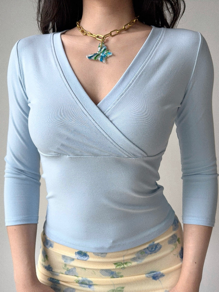 A person wearing a light blue three-quarter sleeve wrap top and a floral skirt, with a gold chain necklace.