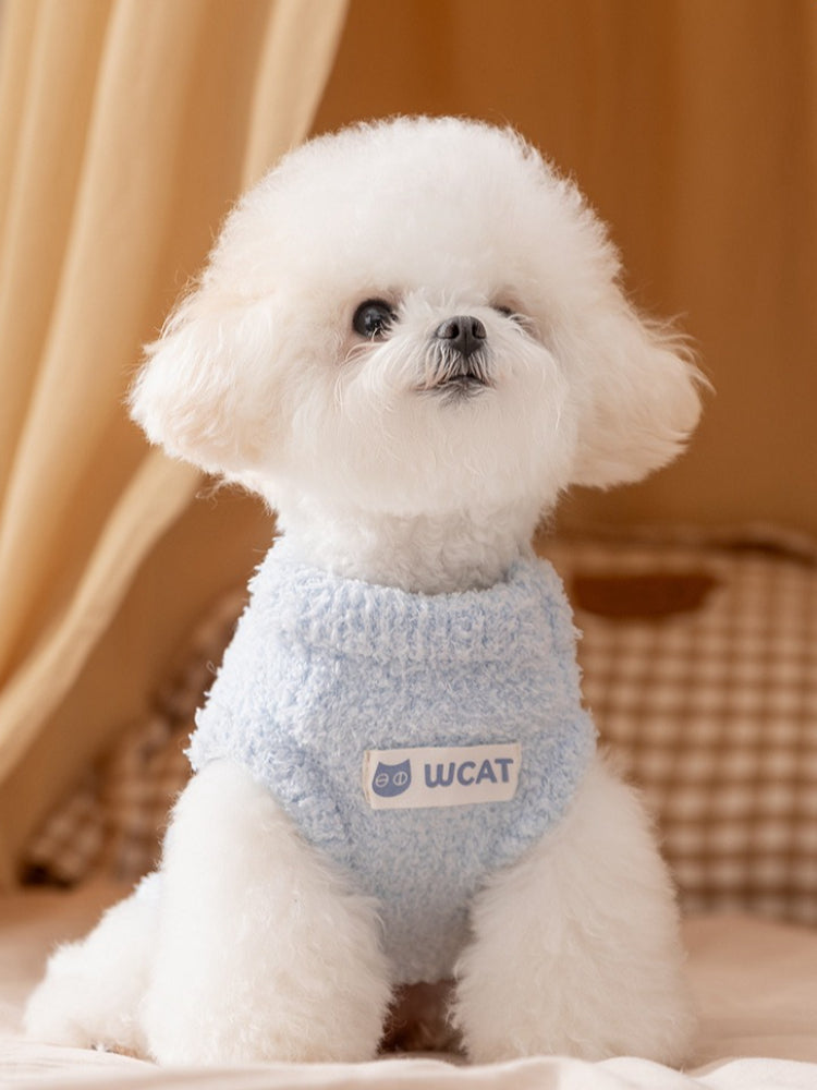 A white bichon frise dog wearing a light blue plush dog sweater with a white 'Wcat' logo patch, sitting on a beige surface.