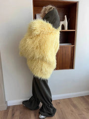 Woman wearing a yellow shaggy faux fur jacket over a dark green hooded sweatshirt with dark green pants.