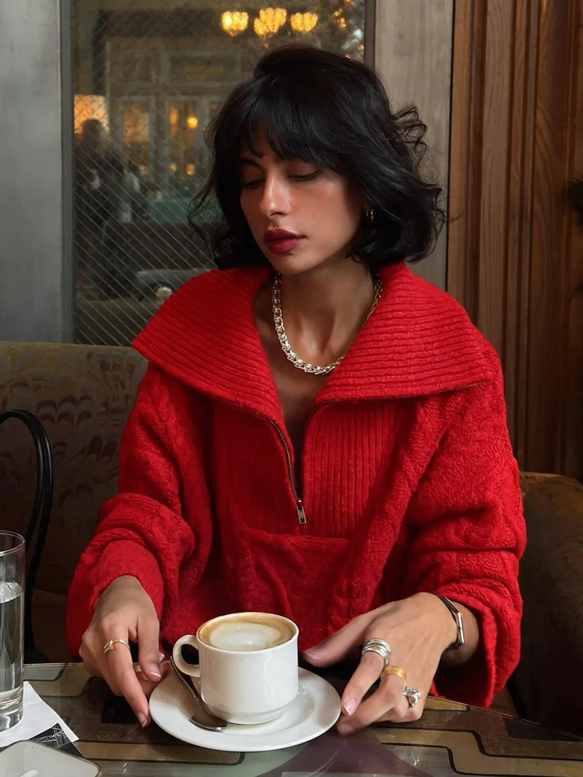 Woman wearing a red textured knit quarter zip sweater with black pants and silver jewelry, holding a coffee cup.
