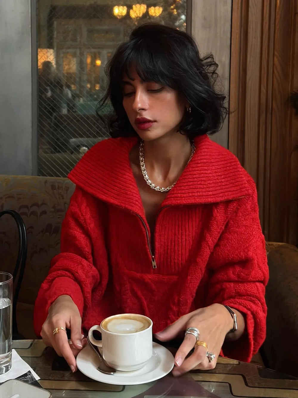 Woman wearing a red textured knit quarter zip sweater with black pants and silver jewelry, holding a coffee cup.