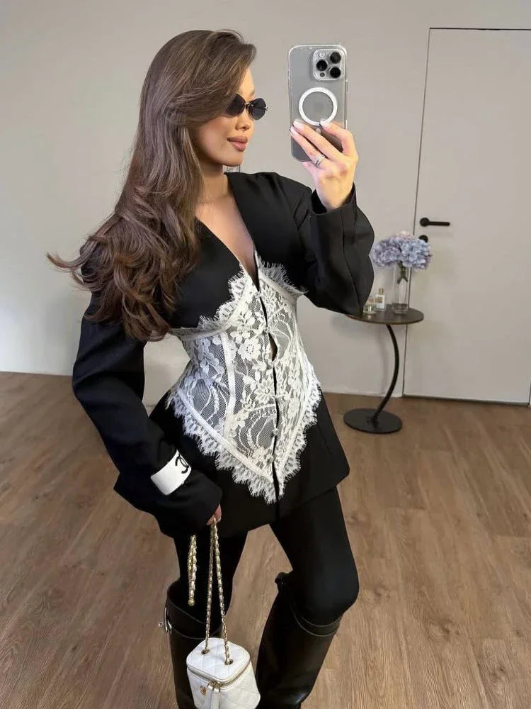 Woman in a black blazer with a white lace corset detail, black leggings, and black boots, holding a white handbag and sunglasses.