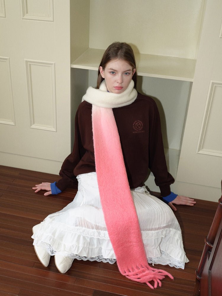 Woman wearing a brown sweatshirt, white ruffled skirt, and a long gradient scarf from cream to pink with fringe detail, sitting on a wooden floor.