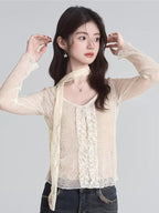 Close-up of a woman's face and upper body wearing a cream lace top with a scarf detail and star earrings.