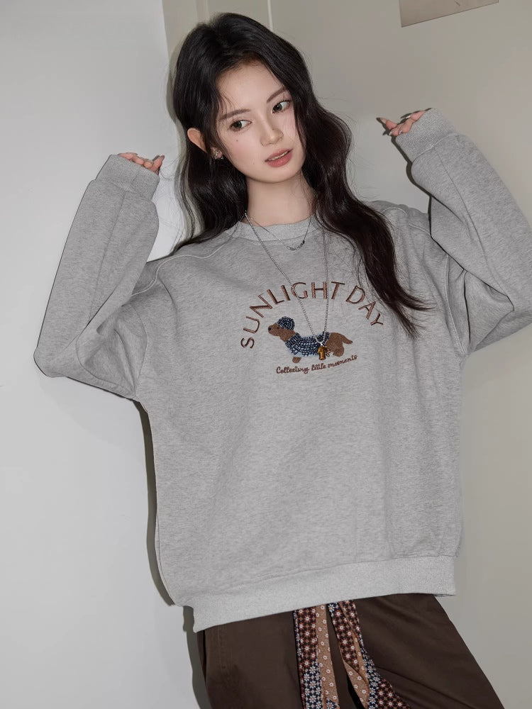 Woman posing with hands raised, wearing a grey crew neck sweatshirt with a dachshund graphic and text "SUNLIGHT DAY".