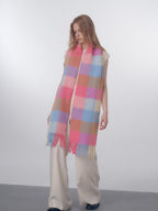 Woman wearing a long, oversized, fuzzy knit scarf with a pink, blue, and tan plaid pattern and fringe details.