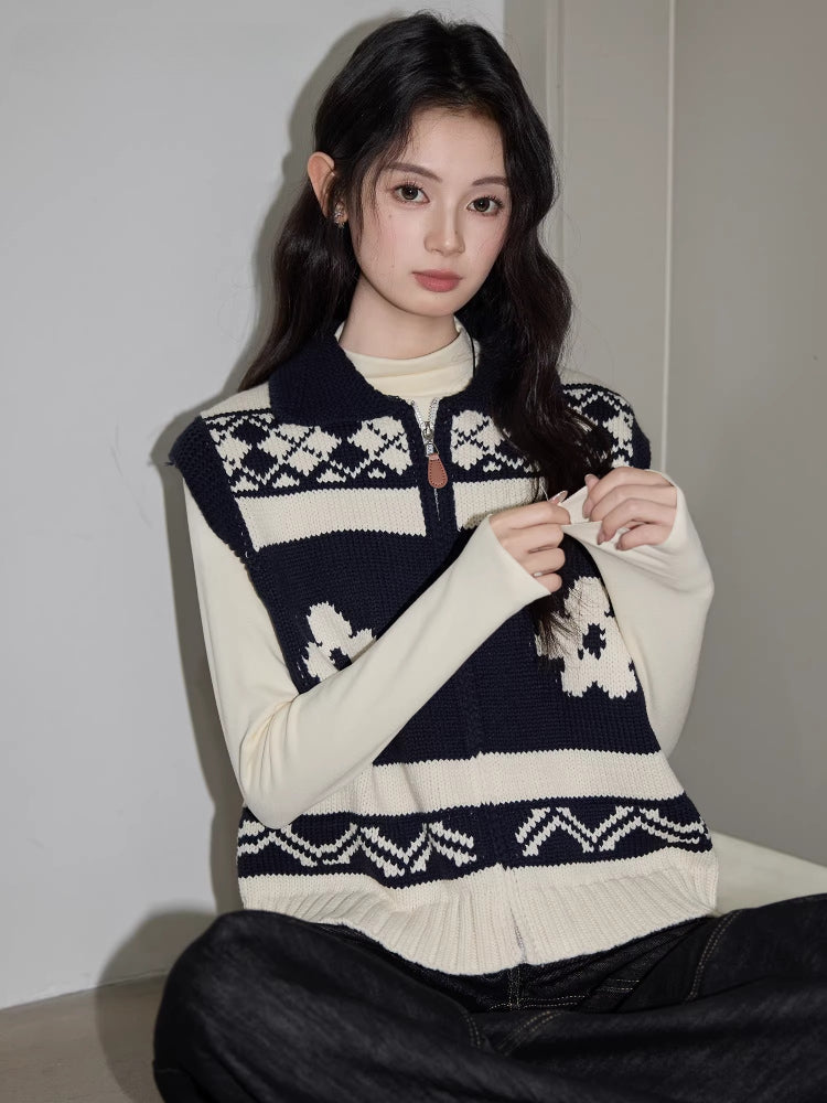 Close-up of a woman wearing a navy and cream floral and geometric knit vest over a cream long-sleeve top and dark wash wide-leg jeans, adjusting the zipper.