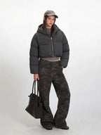 Side profile of a model wearing a cropped grey puffer jacket, grey long-sleeved shirt, and camouflage cargo pants, holding a black tote bag.