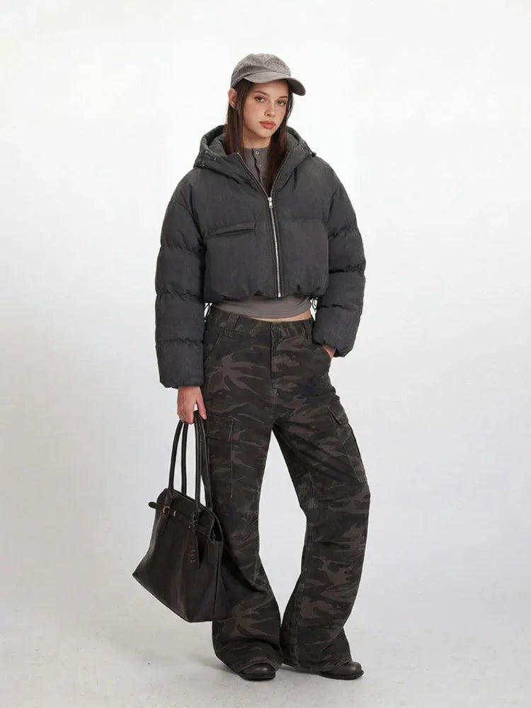 Side profile of a model wearing a cropped grey puffer jacket, grey long-sleeved shirt, and camouflage cargo pants, holding a black tote bag.