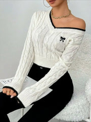Woman sitting with one shoulder exposed, wearing a white cable knit cropped V-neck sweater with black trim and a black bow detail, paired with black pants.