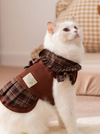 A white cat looking up and to the side while wearing a brown plaid dress.