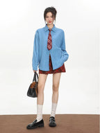 Model wearing an oversized blue collared shirt with a red plaid tie and a red plaid mini skirt, holding a handbag and wearing loafers.