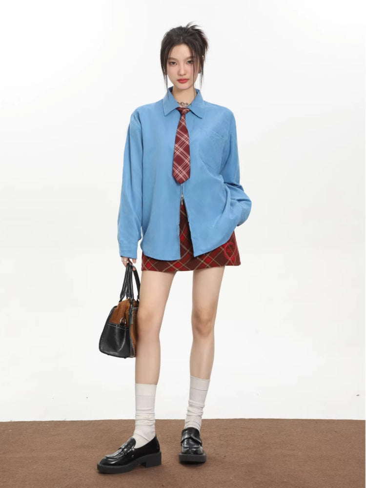 Model wearing an oversized blue collared shirt with a red plaid tie and a red plaid mini skirt, holding a handbag and wearing loafers.