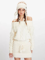 Front close-up of a model wearing a cream off-the-shoulder knit sweater and a matching mini skirt, with an ear flap hat and pink handbag.